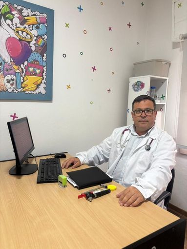 Médico sentado en su escritorio, con bata blanca y estetoscopio. Caricatura en la pared.