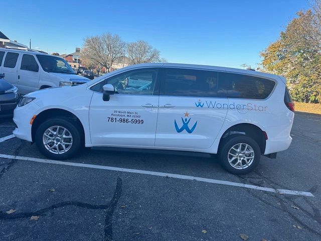 Caregiver in blue scrubs assists elderly person in a pink shirt with walking in a bright room.
