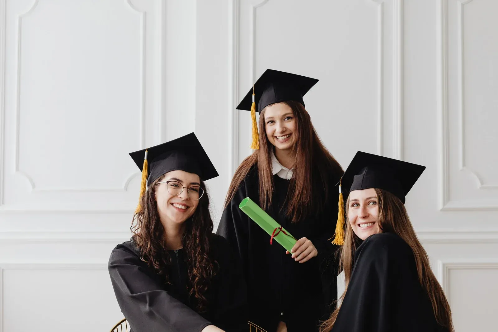 Three graduates in black caps and gowns smiling, one holding a diploma.