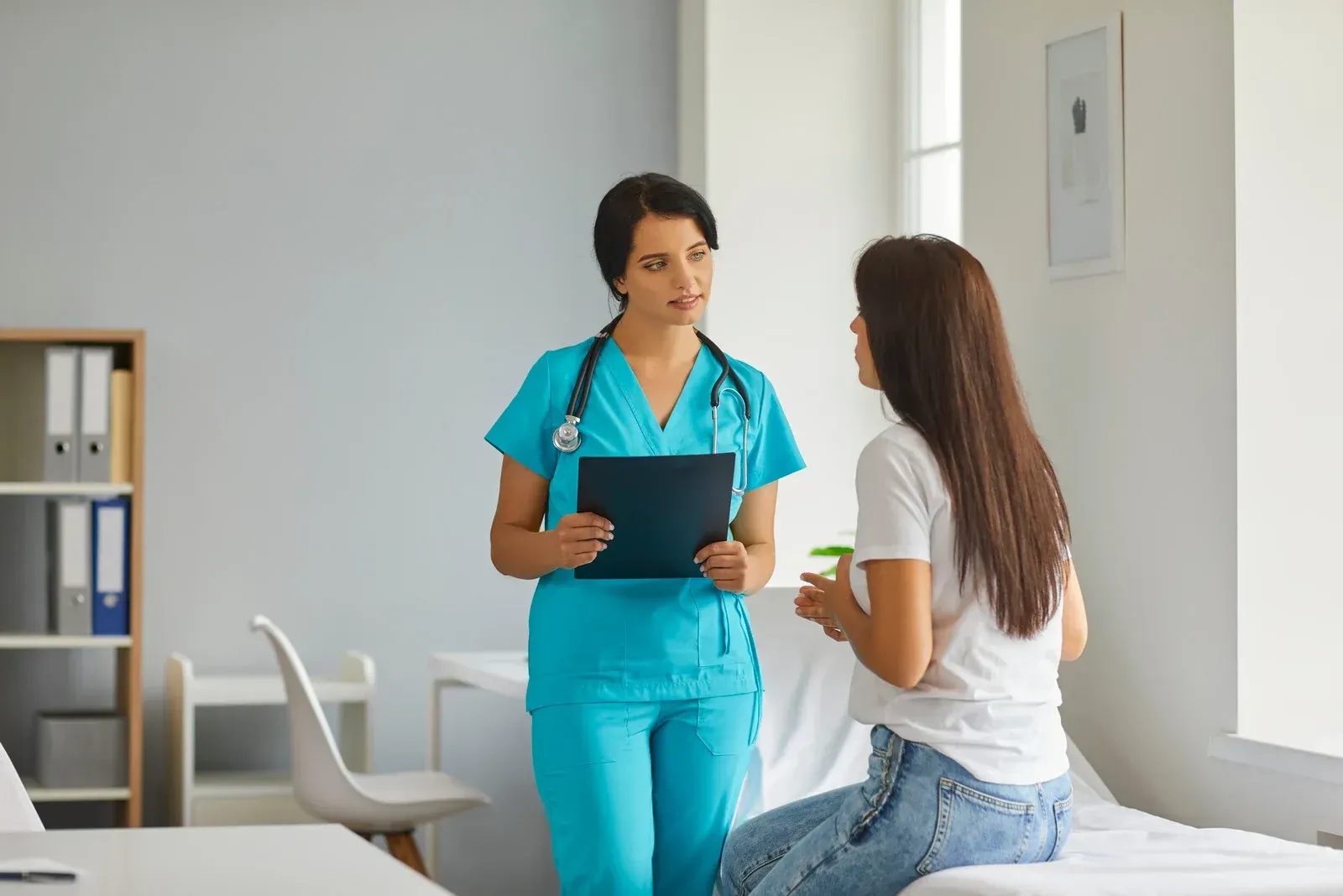 Nurse in blue scrubs consulting a patient seated on a medical exam table, holding a clipboard in a medical office.