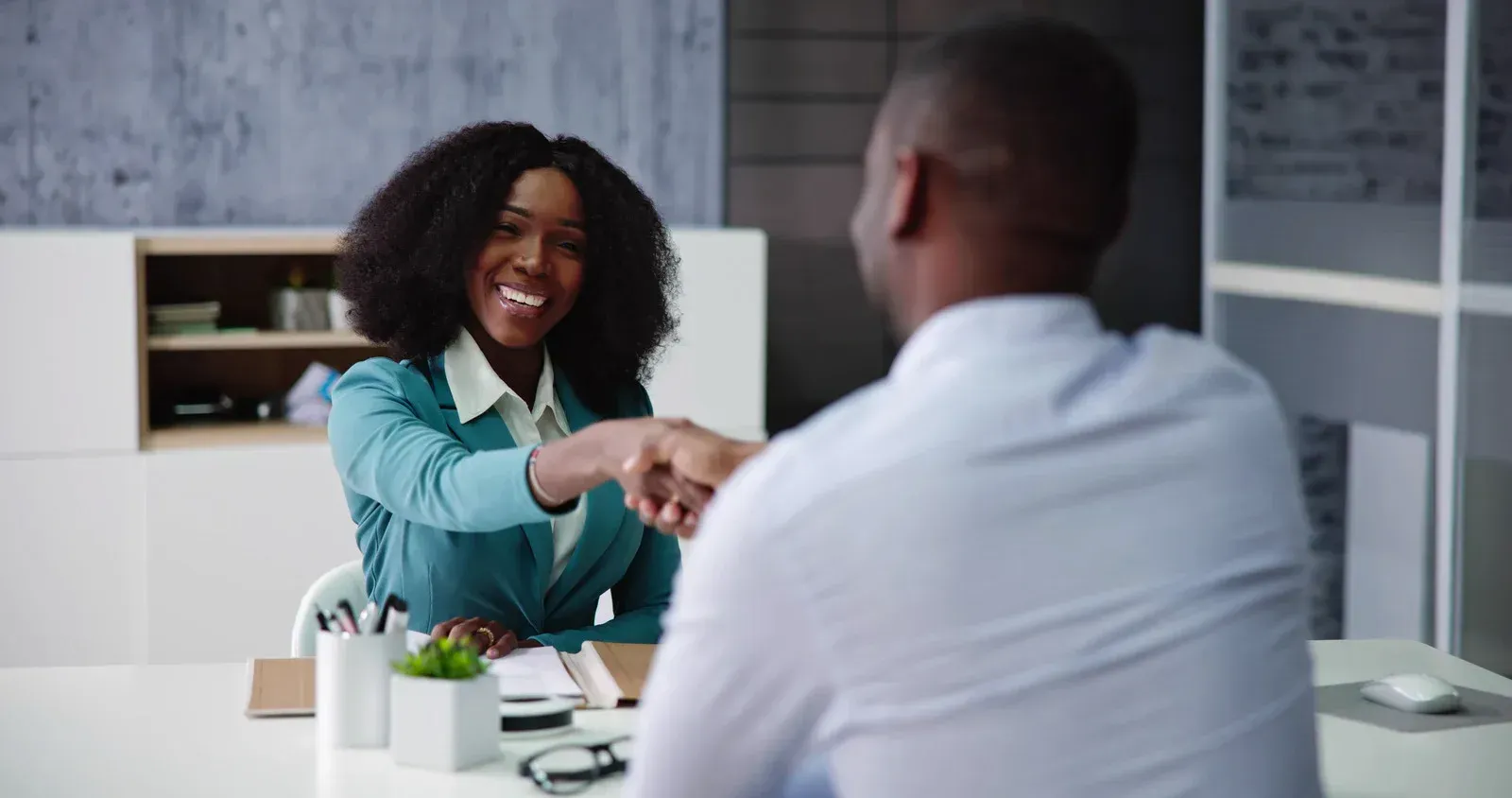 Woman in teal blazer shaking hands with a person, smiling in an office setting.