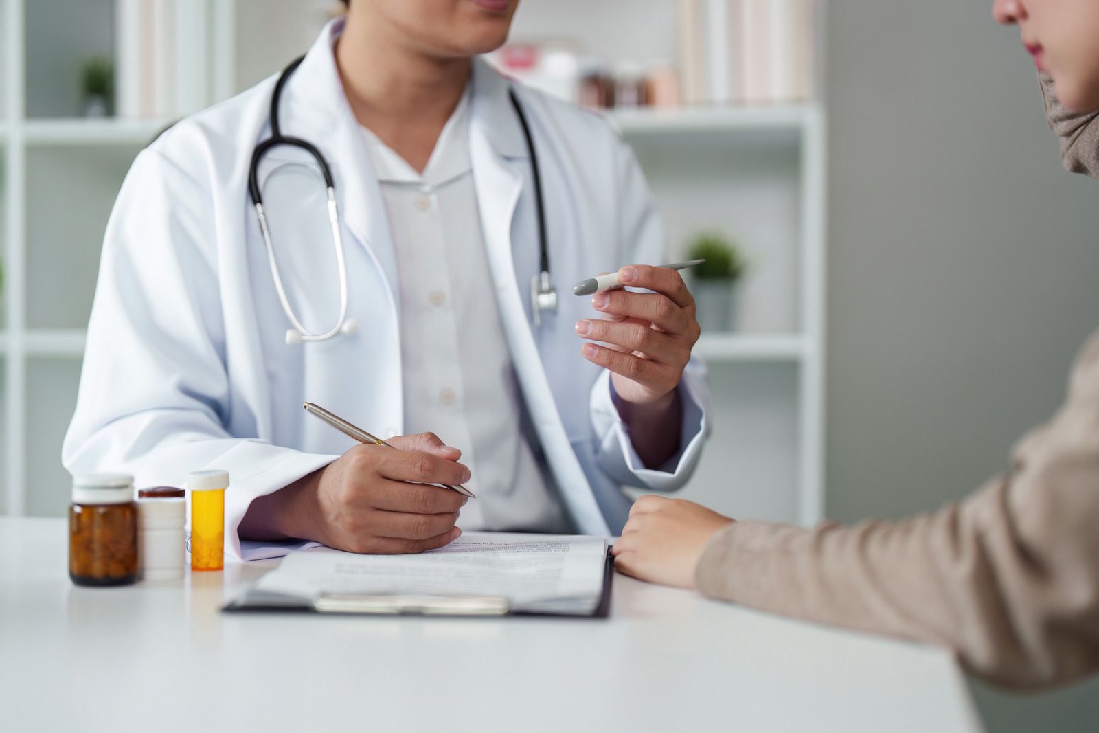 Doctor in white coat reviewing paperwork with a patient at a desk. Bottles of medicine nearby.
