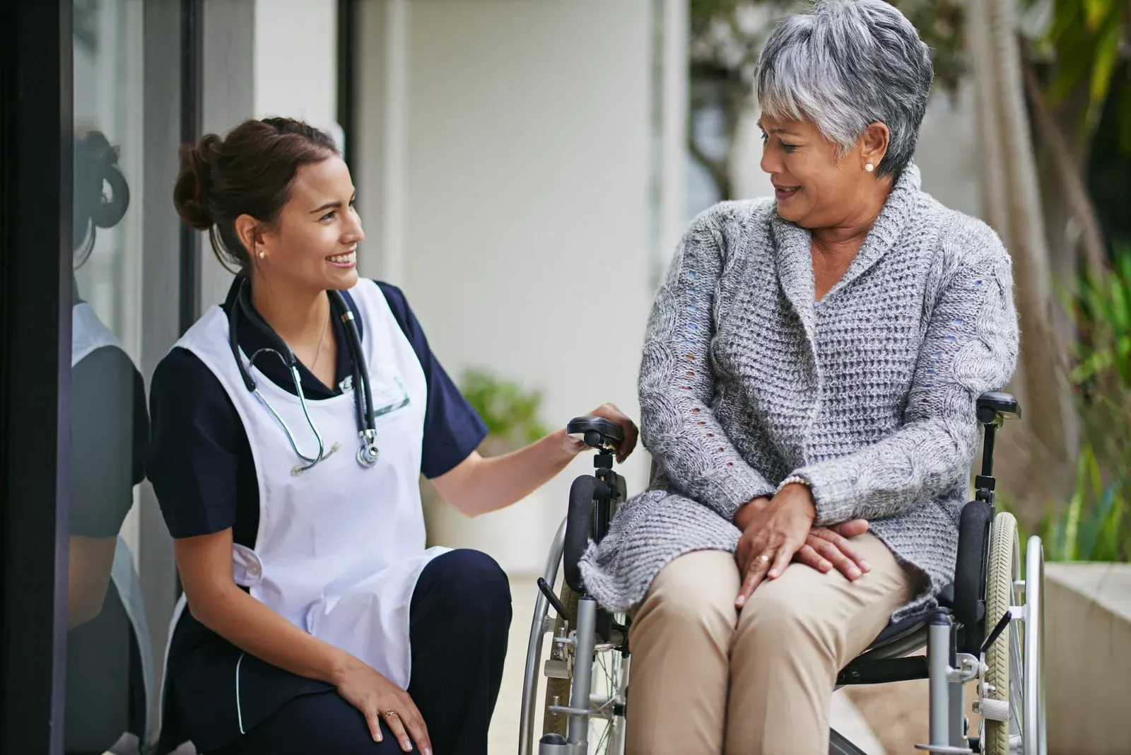 Caregiver with stethoscope smiles at an elderly person in a wheelchair outside.