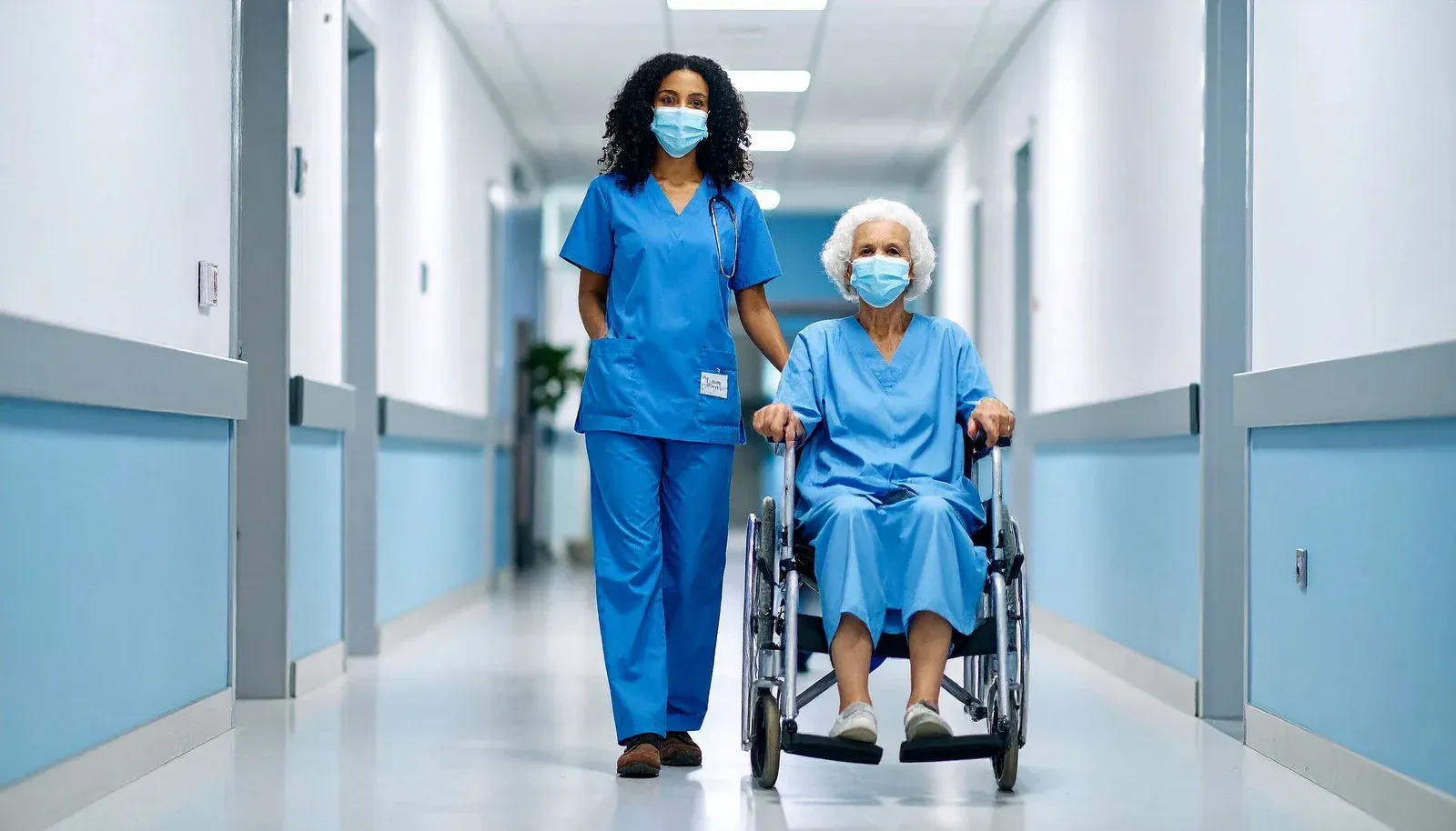 Nurse in blue scrubs pushes a person in a wheelchair down a hospital hallway. Both are wearing face masks.