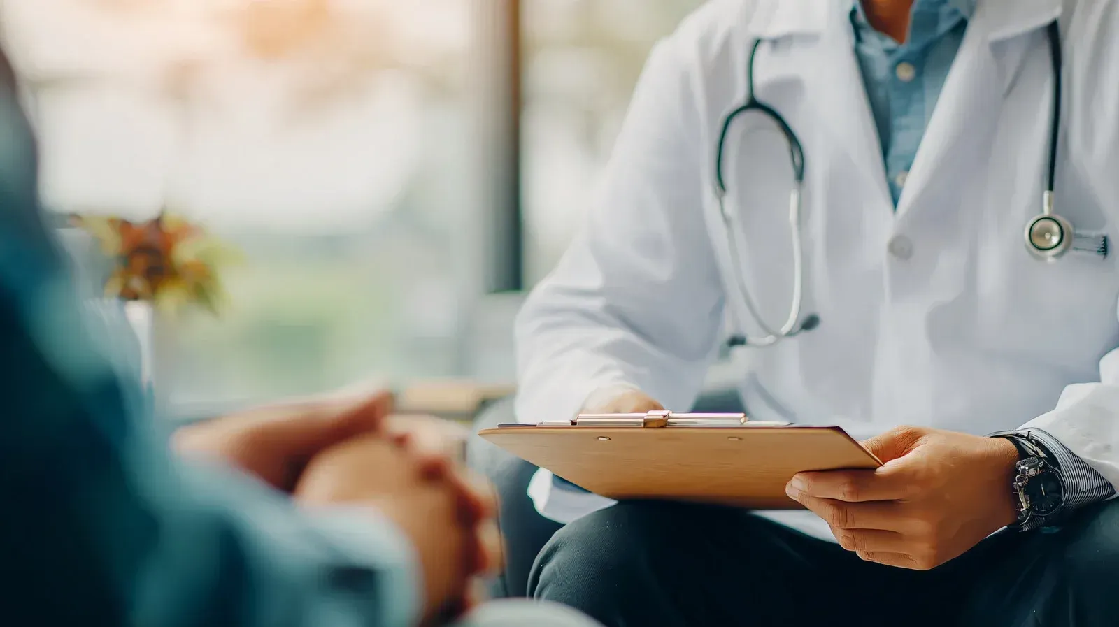 Doctor in white coat with stethoscope, holding a clipboard, speaking to a patient.