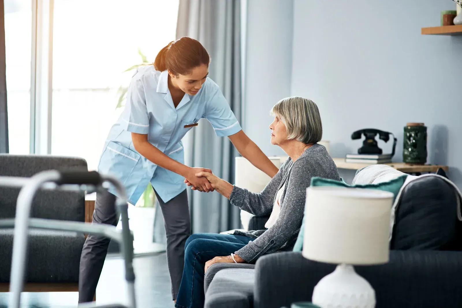 Caregiver helping senior woman sit on sofa indoors.