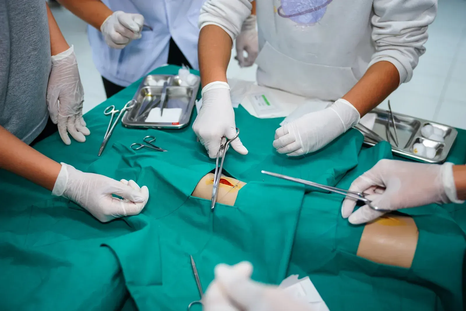 Hands wearing gloves performing surgery with surgical instruments on a green draped surface.