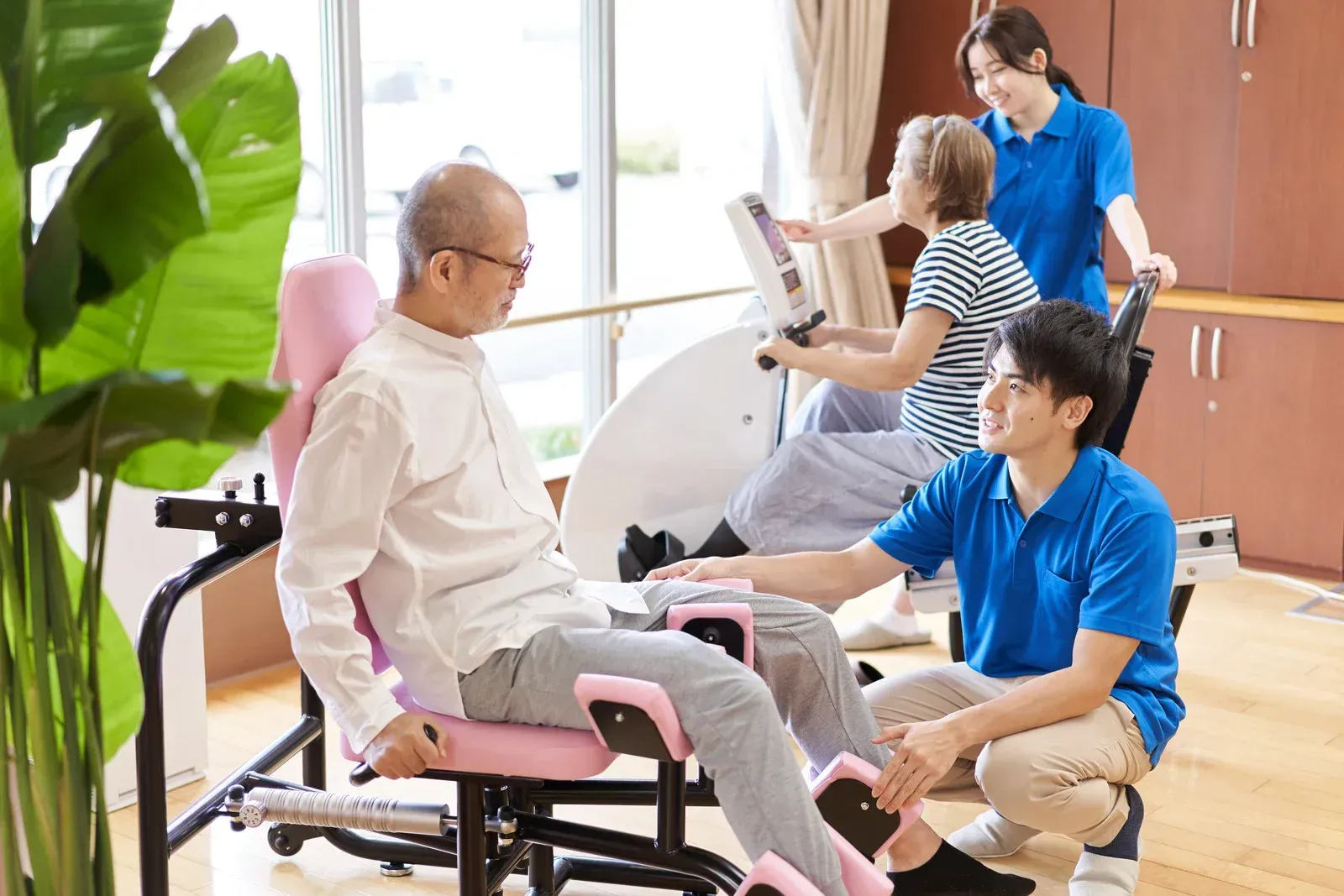 Two people in therapy, one using a leg exercise machine. Two assistants observe and help.