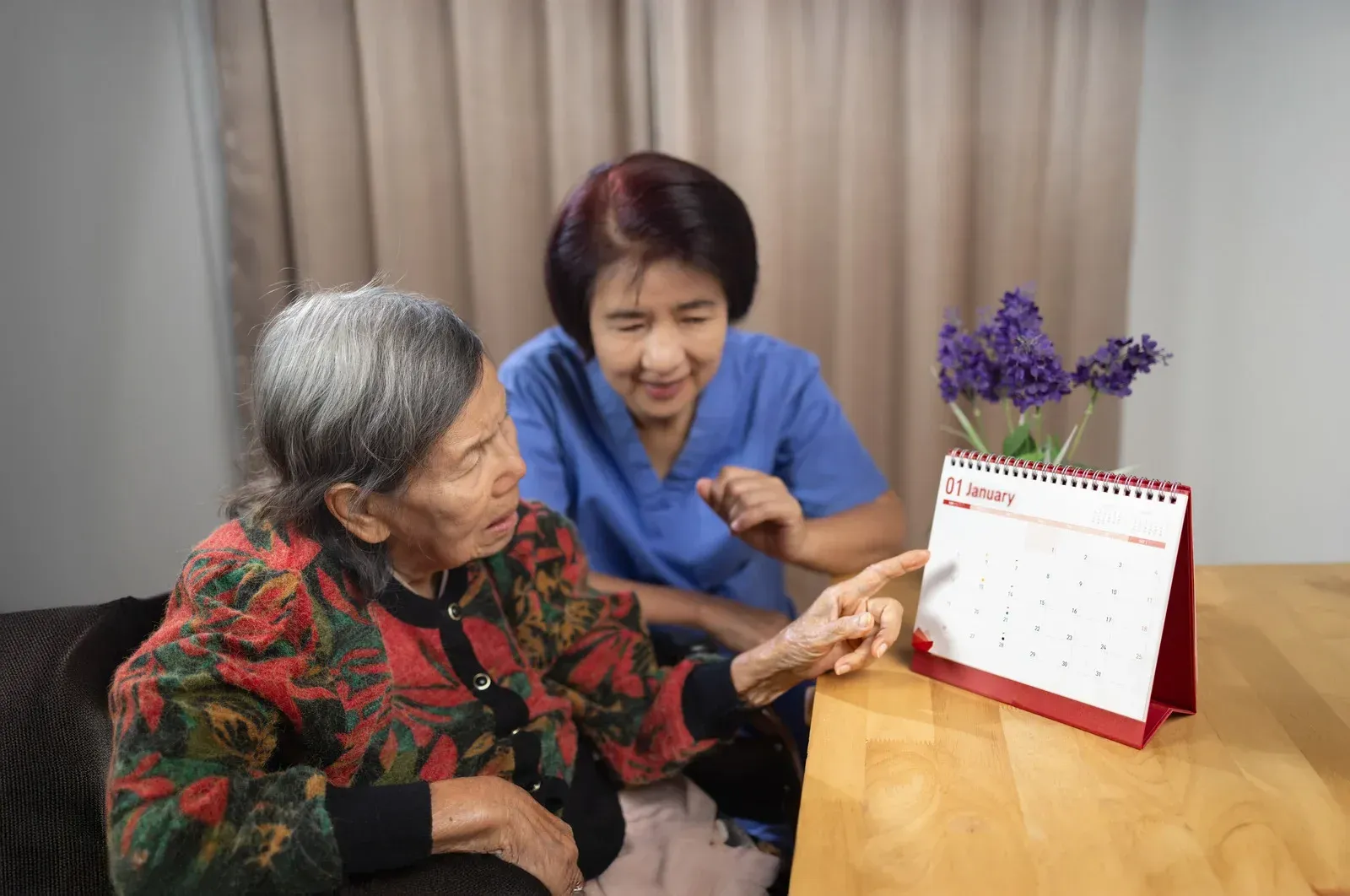 An older person and a caregiver looking at a calendar on a table in a room with purple flowers.