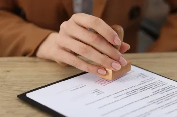 Person stamping a document on a desk; close-up of hand holding a wooden stamp, brown jacket.