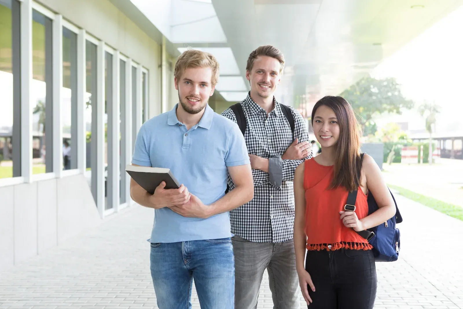 Three students smiling outside a building, one holding a book, one with a backpack.