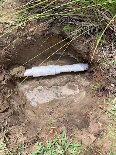 White PVC pipe exposed in a hole in the ground, surrounded by dirt and grass.