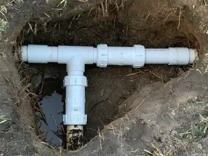 White PVC pipes in an excavated dirt hole. A T-fitting joins two horizontal pipes with a vertical pipe, surrounded by mud.