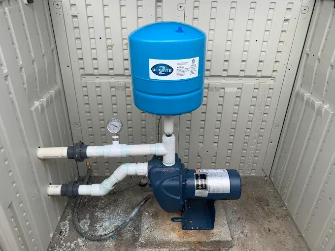 Blue water pump with tank, gauges, and PVC piping inside a small beige shed.