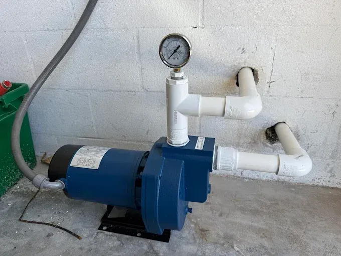 Blue water pump with pressure gauge and white PVC pipes on a white wall.