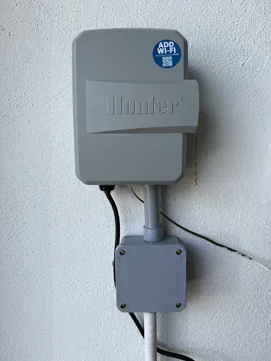 Gray Hunter irrigation controller mounted on a white wall, with electrical conduit and wiring visible.