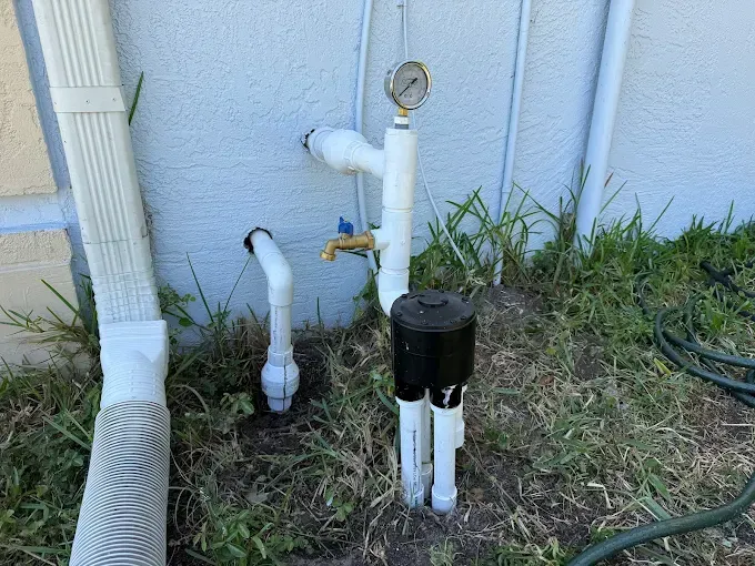 Plumbing pipes and a gauge on a beige wall, surrounded by grass. A rain gutter is on the left.