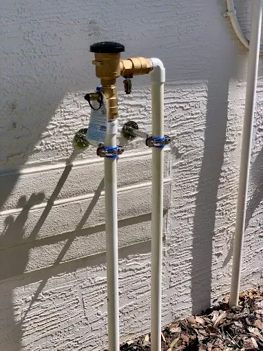 Outdoor irrigation system with pipes, valves, and a backflow preventer against a textured wall.