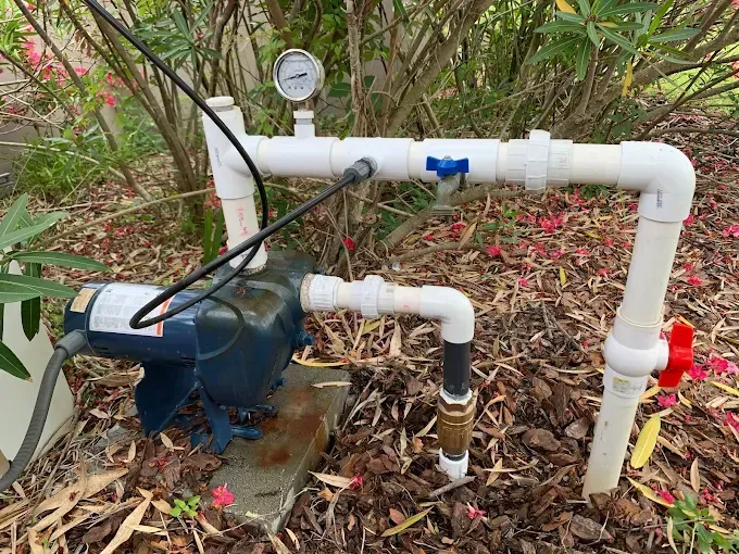 Blue water pump with white pipes, gauge, and red valve in a garden setting.