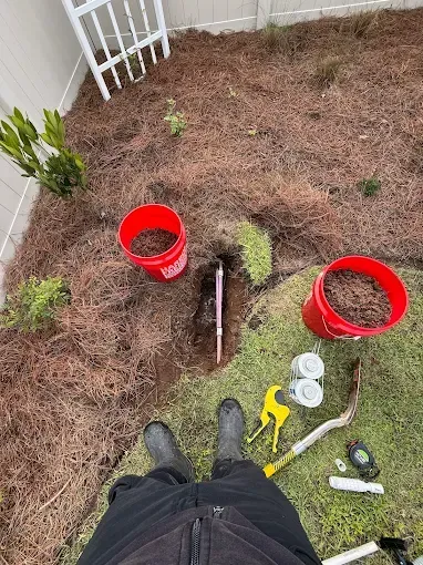 Person in boots standing over a garden bed with a dug hole, buckets, and tools.