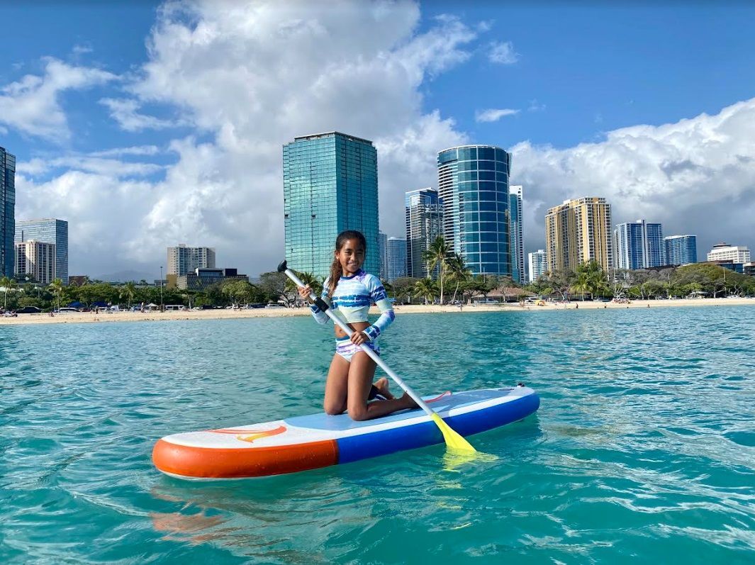 Young girl on a paddle board on the ocean with buildings in the background. She goes to the best pediatrician in Hawaii.