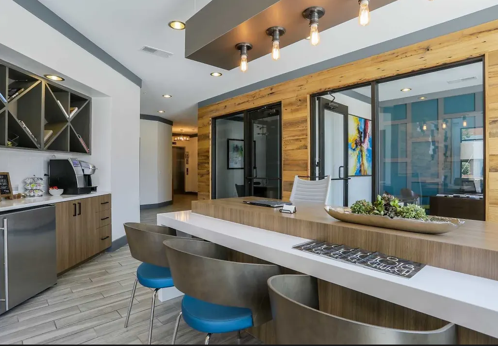 People working at a large wooden table in a bright office; two using laptops, one writing, plants and supplies on the table.