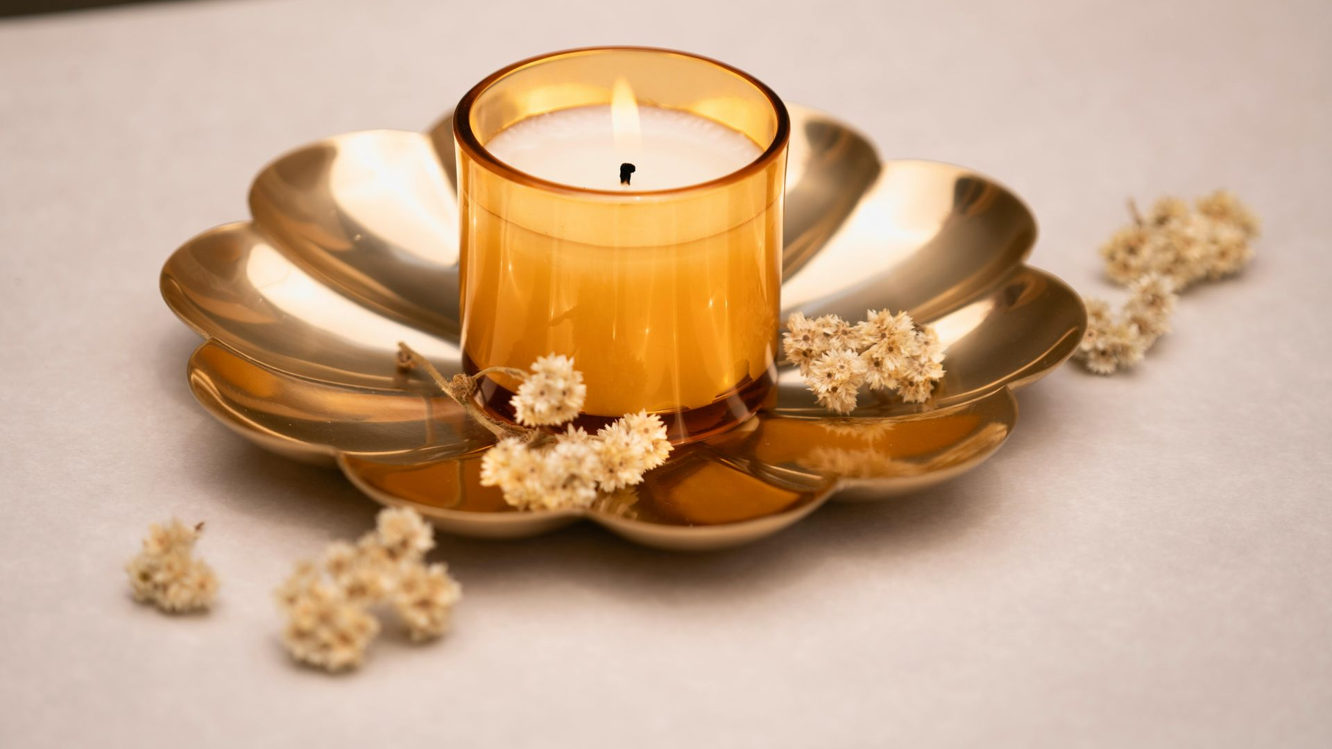 Lit candle in a gold glass holder on a petal-shaped gold dish, with dried white flowers.