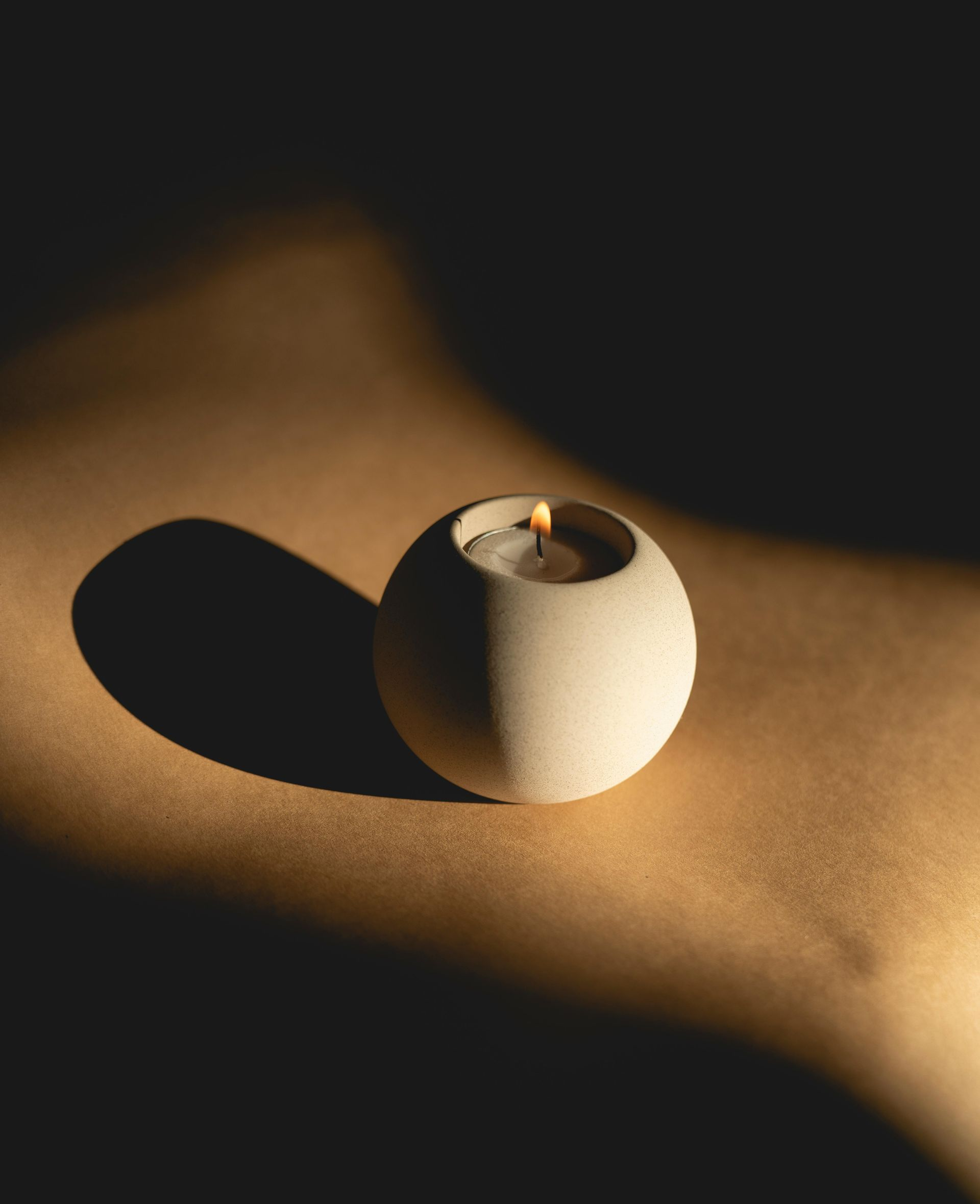 Lit candle in a spherical, light-colored holder casting a shadow on a brown surface. Dark background.