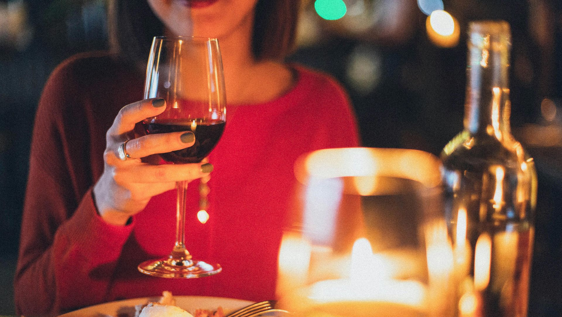 Woman in red sweater holds glass of red wine at candlelit dinner, bottle on table.