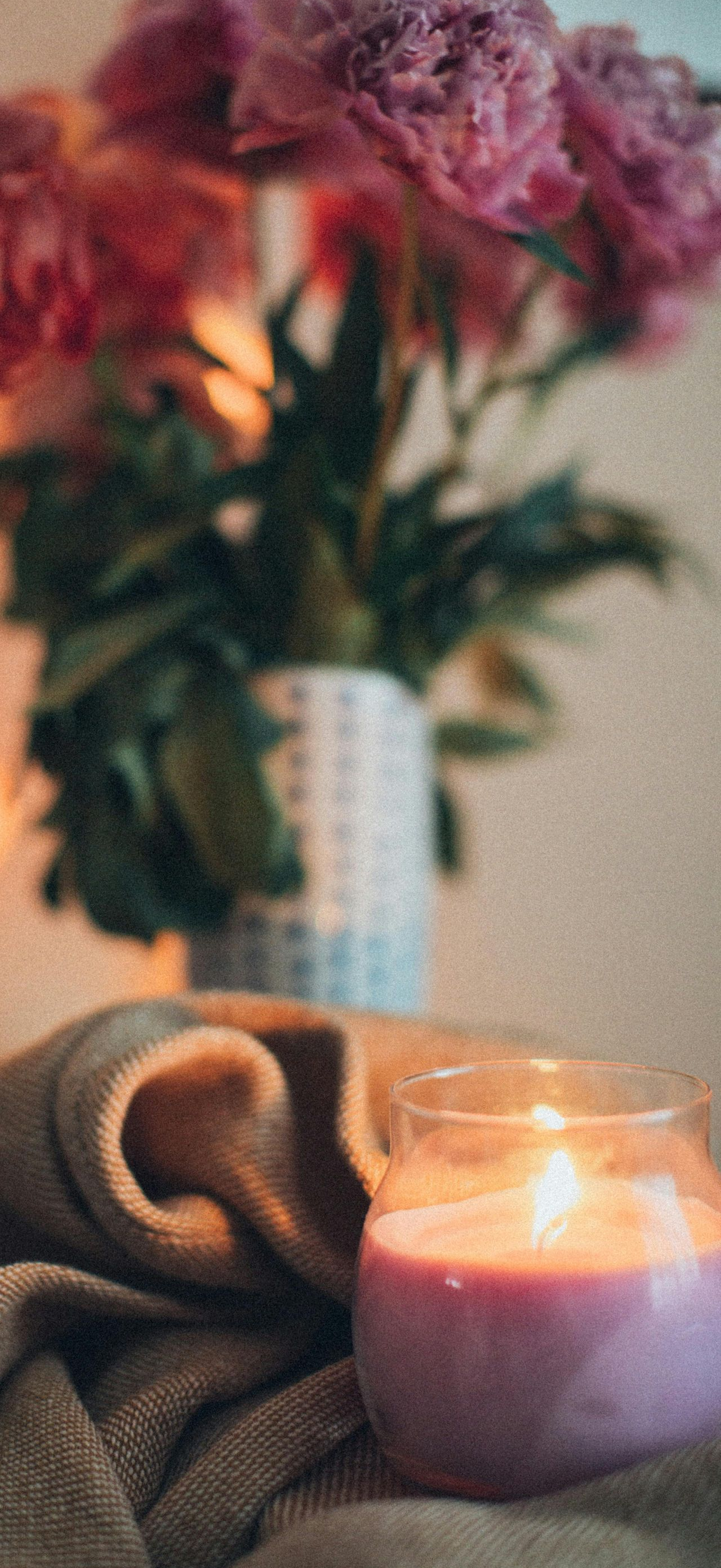 Pink flowers in vase, lit candle, and blanket on a table. Warm, soft lighting.
