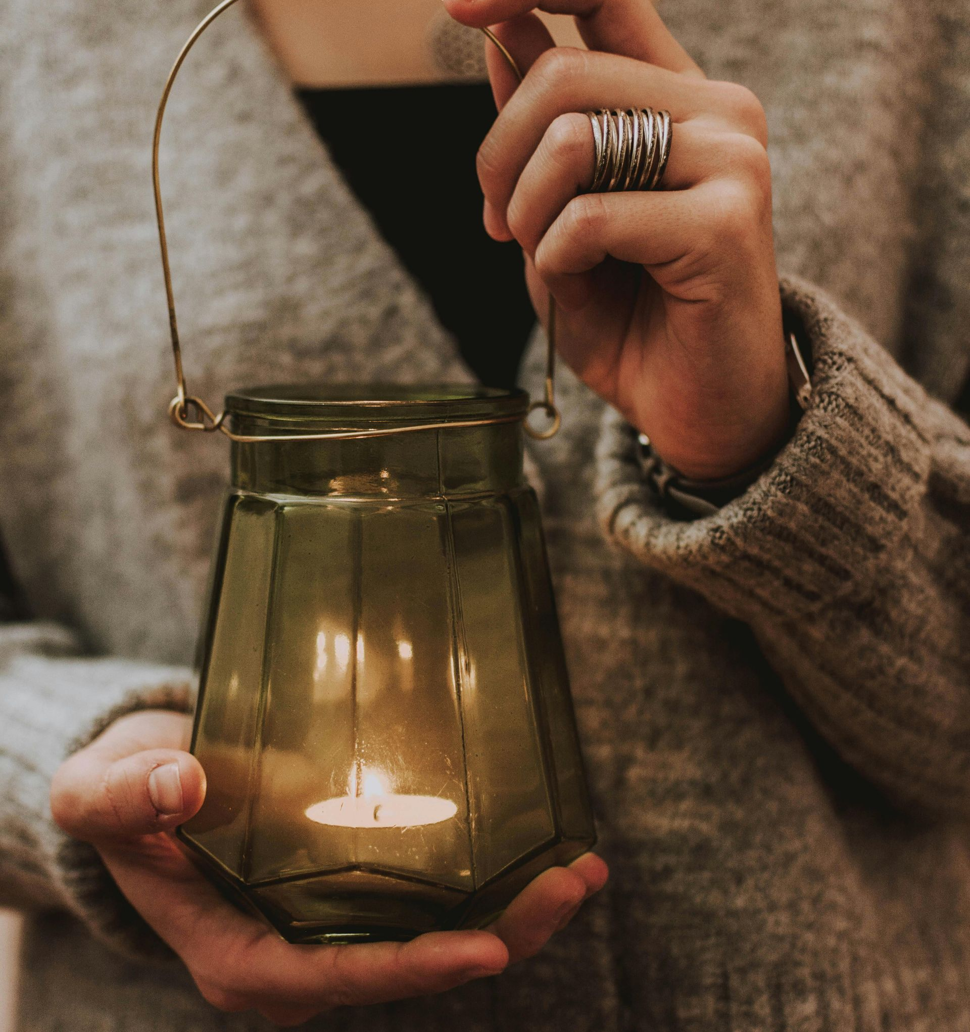 Person holding a green glass lantern with a lit candle, wearing a gray sweater.