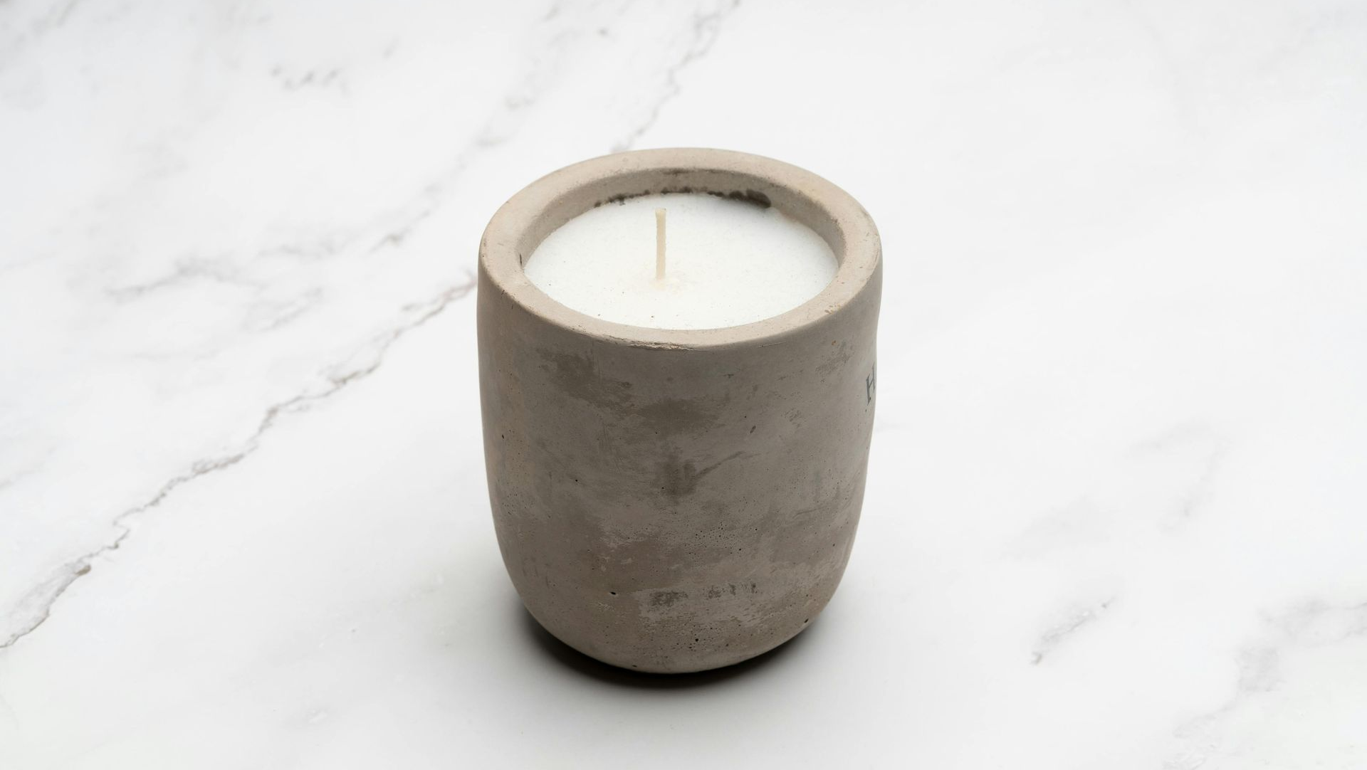 Candle in a cylindrical concrete holder, white wax, single wick, on a white marble surface.