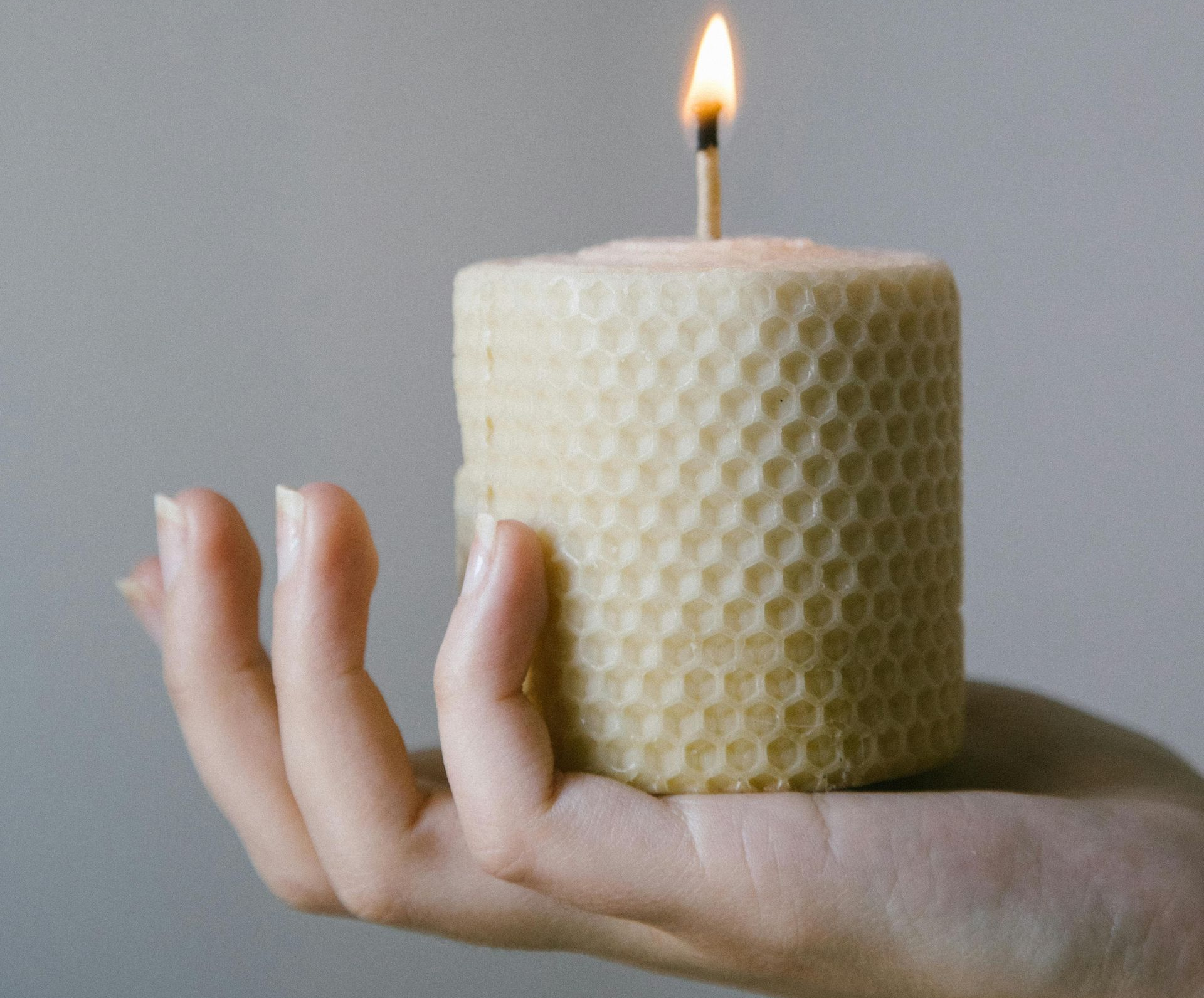 Hand holding a lit, cylindrical beeswax candle with a honeycomb pattern.
