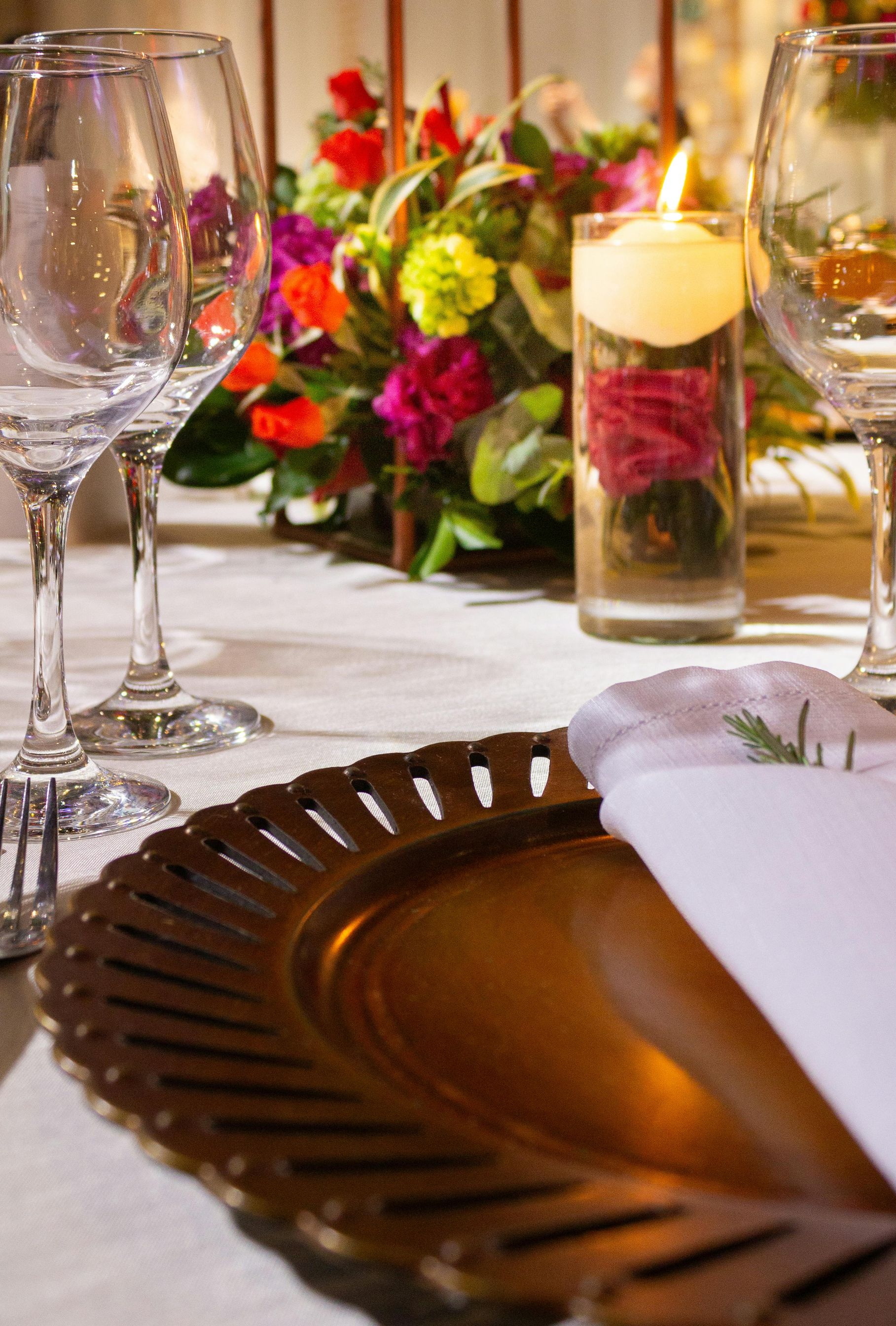 Formal table setting with gold charger plate, glassware, flowers, and a candle.