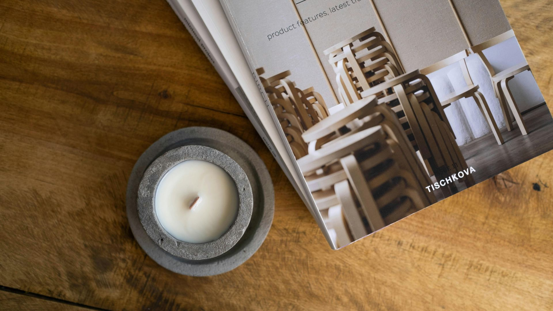 Candle in gray holder next to a stack of catalogs on a wooden surface.