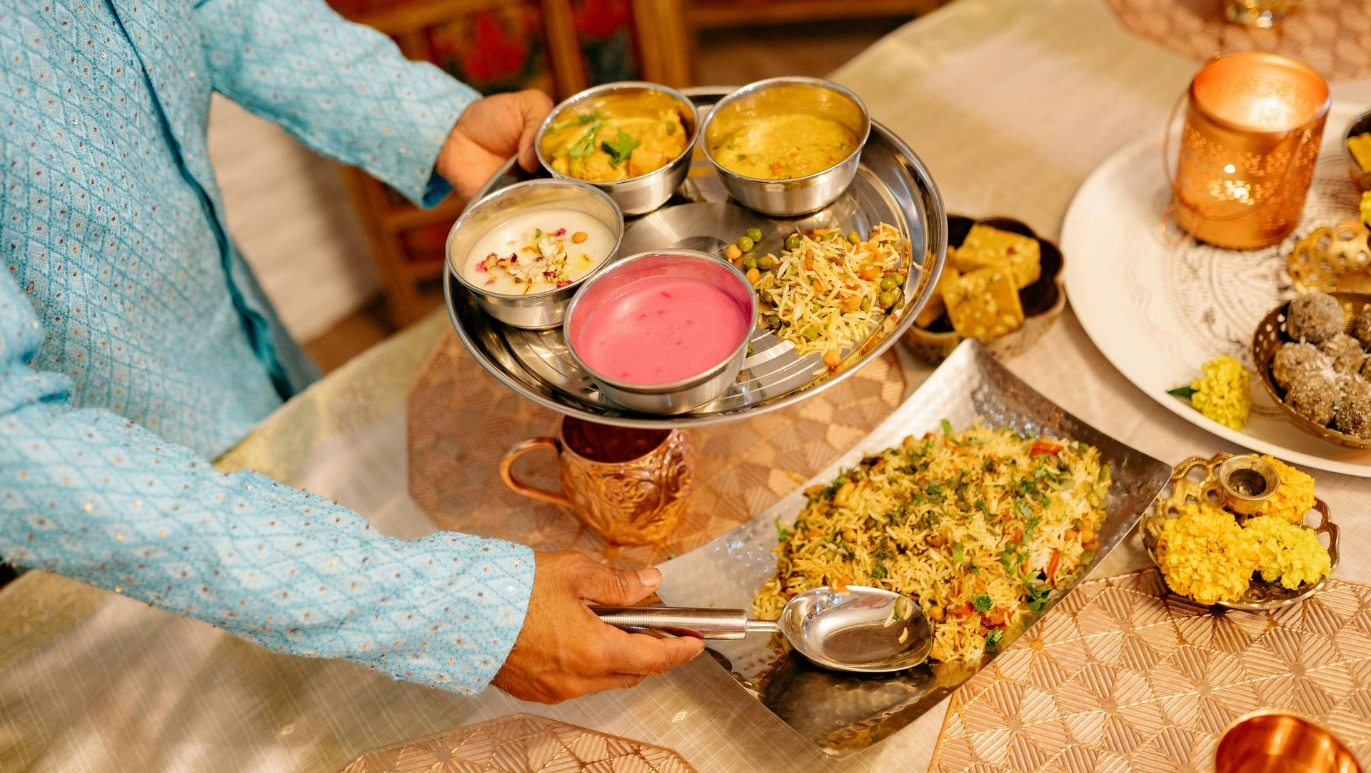 Person holding a silver tray with various Indian dishes at a decorated table, lit by a candle.