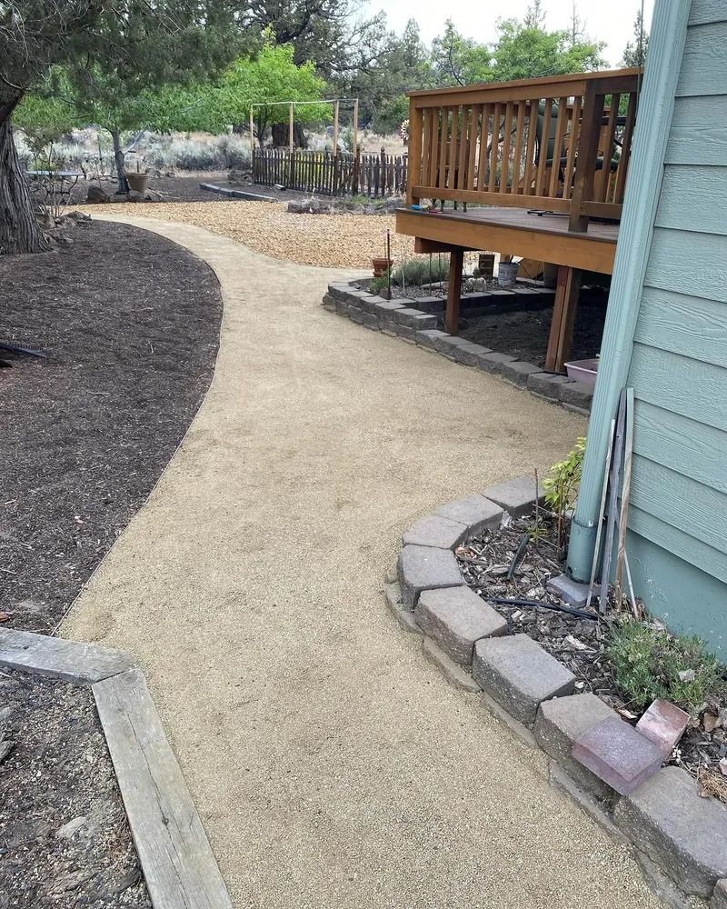 A gravel pathway curves through a backyard, bordered by mulch and a deck.