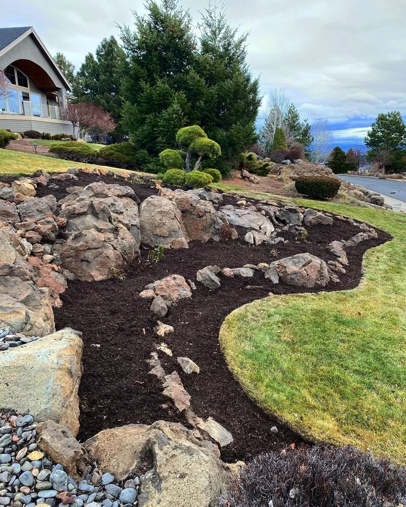 Landscaped hillside with large rocks, dark mulch, and green grass.