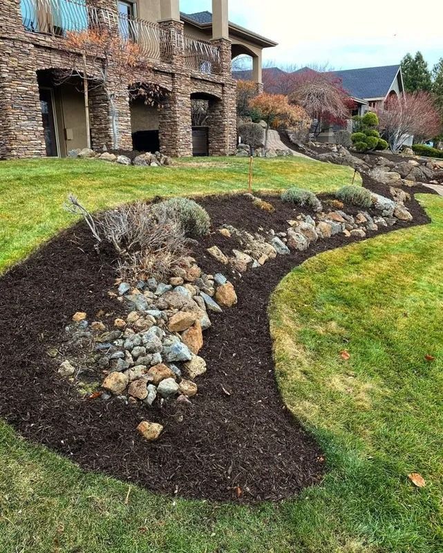 Curved mulch bed with rocks and plants in front of a stone house with green lawn.