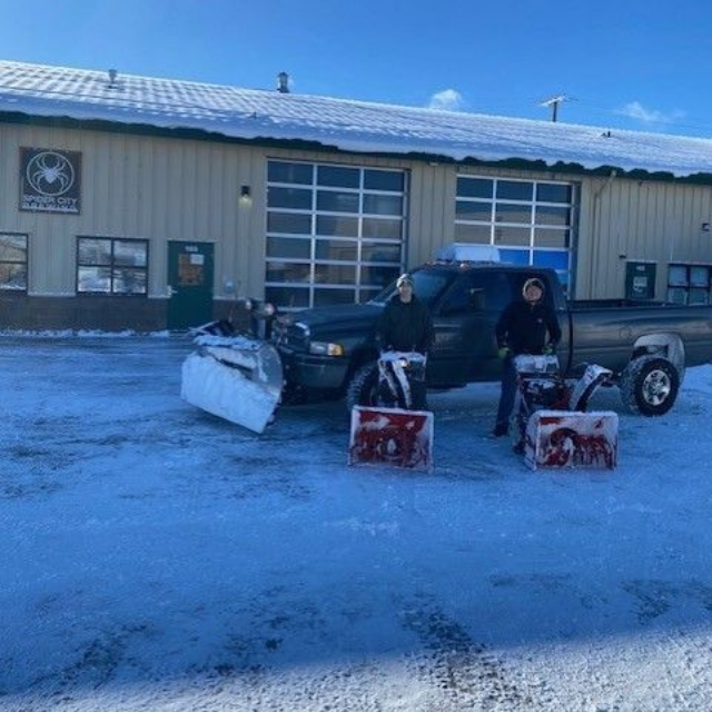 Two people standing by a truck with a snowplow in front of a building covered in snow. Snowblowers on ground.
