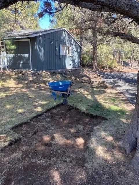 Yard with dug-up dirt patch, wheelbarrow, and small blue building in the background. Sunny day, shadows.