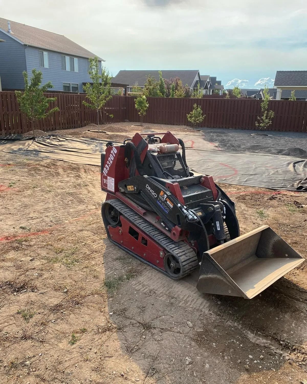 Red mini skid steer loader with bucket on a dirt yard, fence and houses in background.