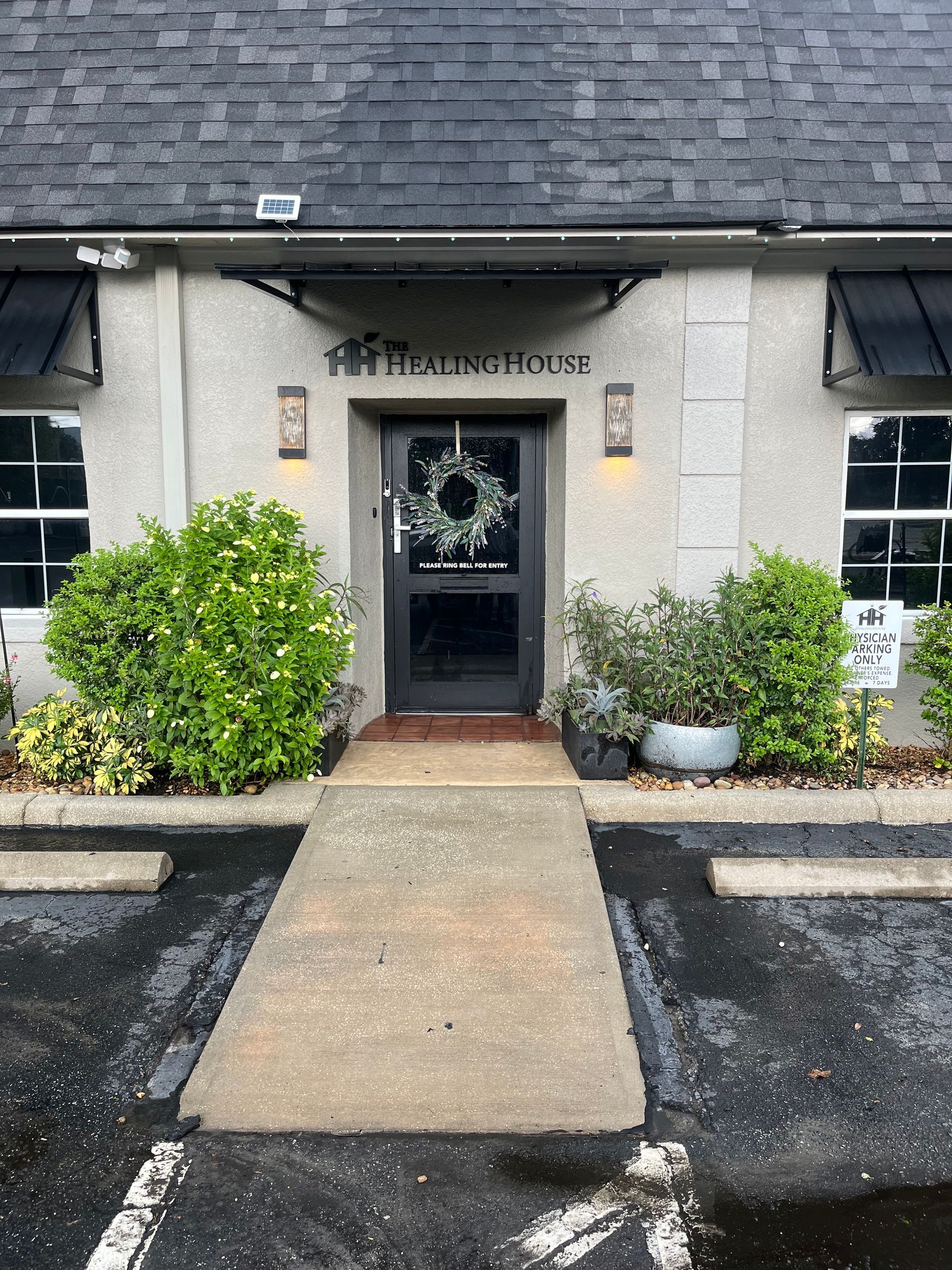 A concrete ramp leads to a building with a dark door and a wreath, flanked by shrubs and windows.