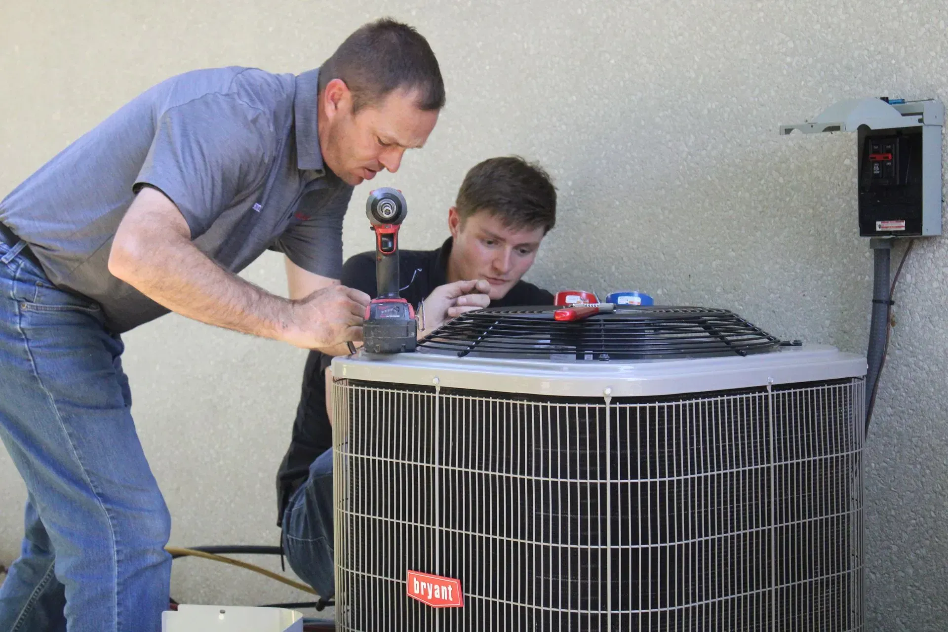 Two men are working on an air conditioner outside of a building.