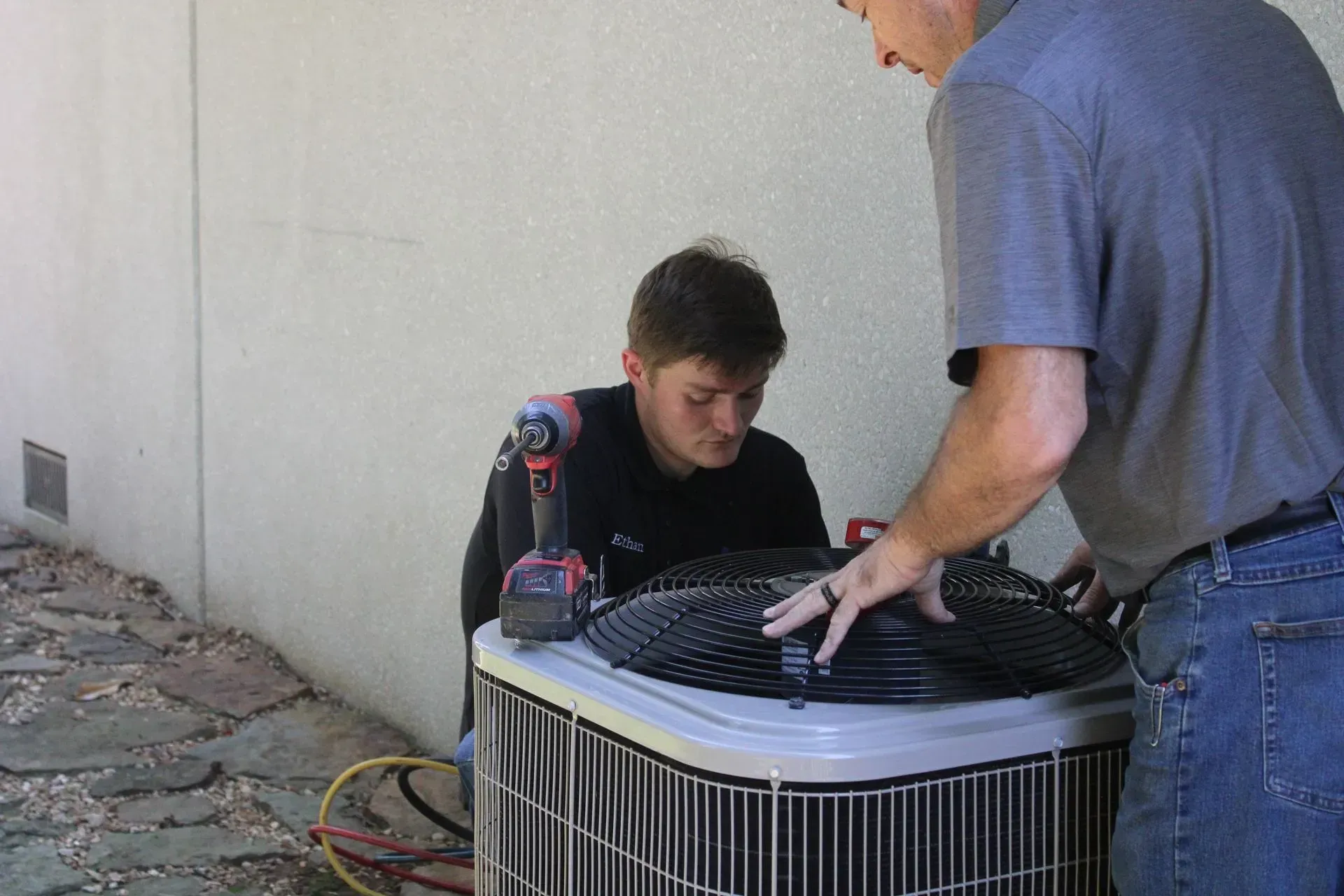 Two men are working on an air conditioner outside of a building.