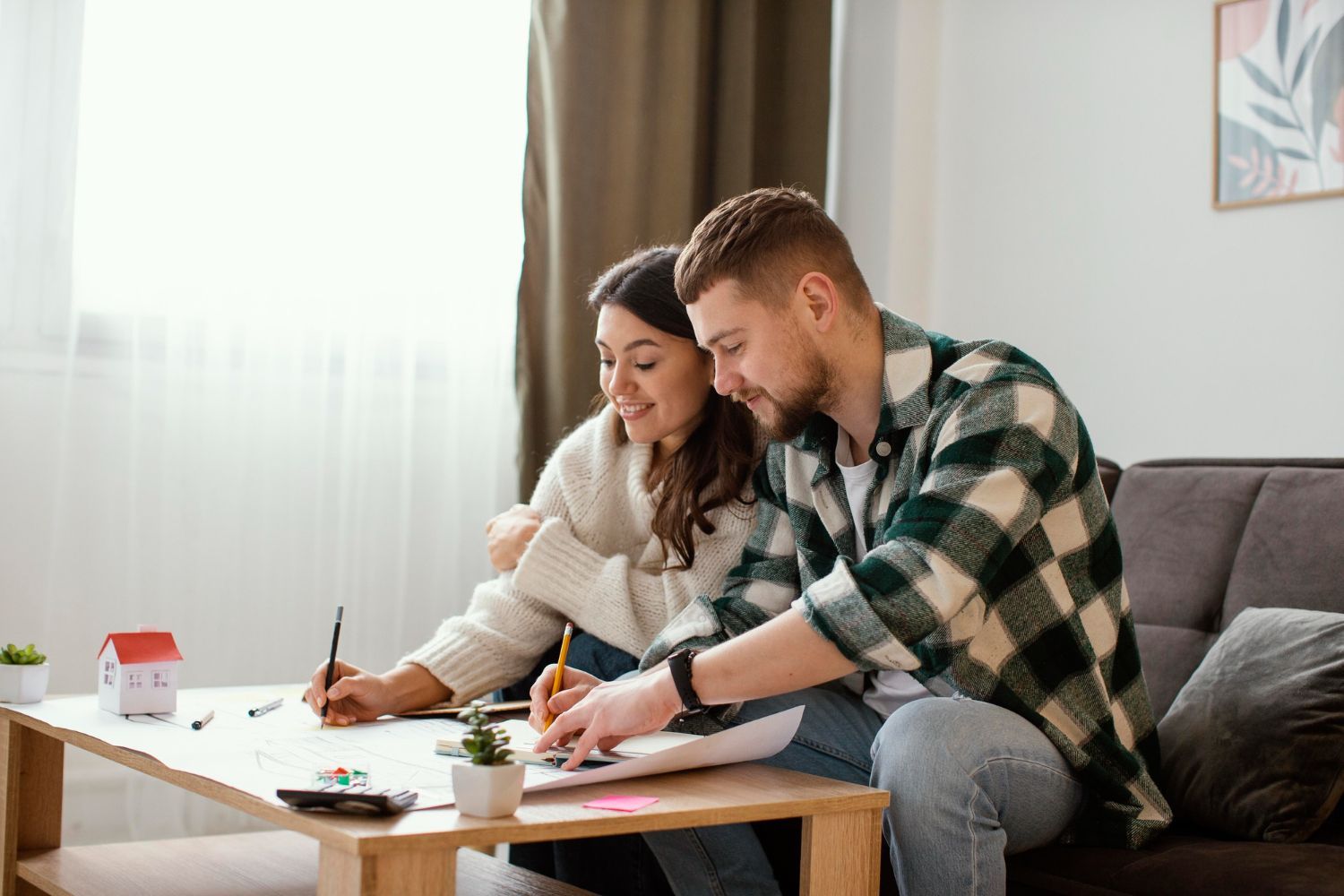 Couple seated on couch, drawing plans at a table, a model house on the table.