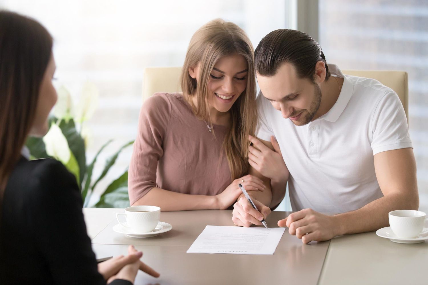 Couple signing documents at a table with a businesswoman. Smiles, coffee cups, and office setting.
