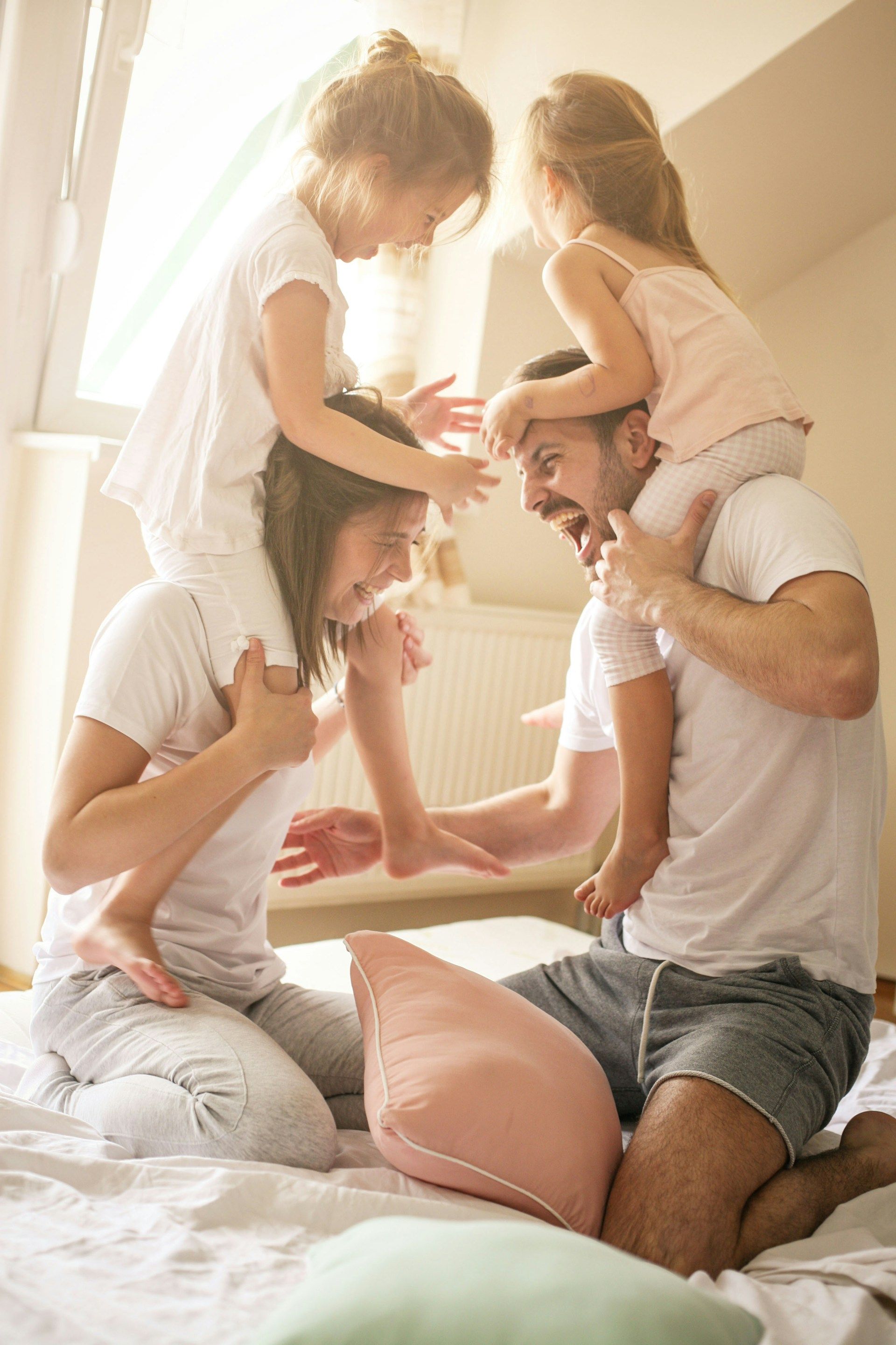 Family playing on bed: parents with daughters on their shoulders, laughing, sunny room.