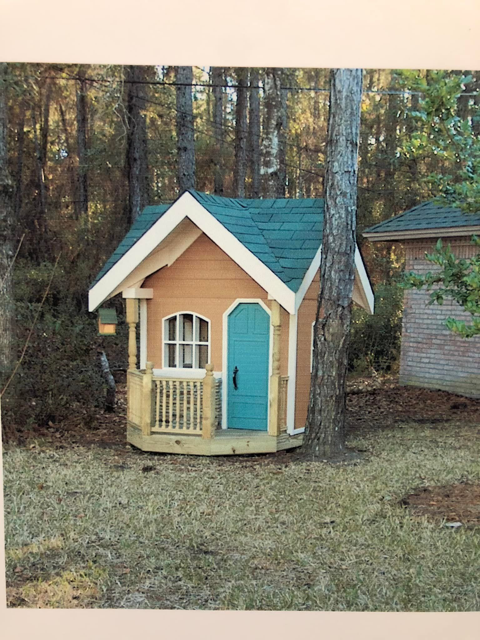 Small wooden playhouse with teal door and green roof, in a grassy yard surrounded by trees.