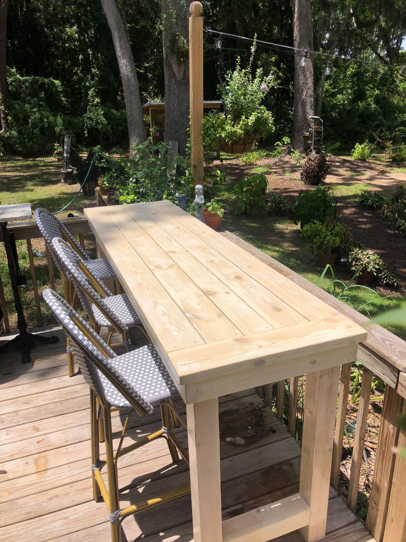 A wooden outdoor bar with light-colored top, wooden legs, and gray-patterned chairs on a deck with trees in the background.